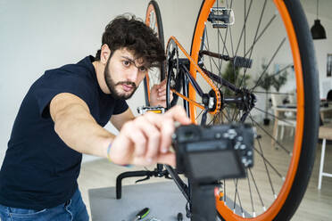 Junger männlicher Vlogger, der den Blick auf sein Smartphone einstellt, während er eine Anleitung zur Reparatur eines Fahrrads zu Hause filmt - GIOF11966