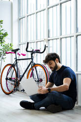 Man using laptop while sitting near window by bicycles at home - GIOF11970