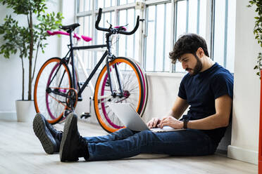 Entspannter Mann, der einen Laptop benutzt, während er in der Nähe von Fahrrädern zu Hause sitzt - GIOF11971