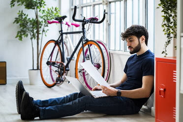 Young man holding document while using laptop by bicycles at home - GIOF11973