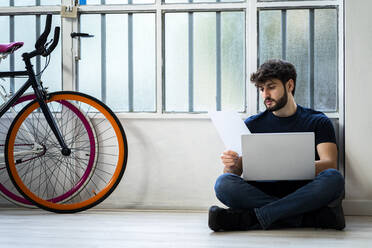 Man holding document using laptop while sitting near window at home - GIOF11974