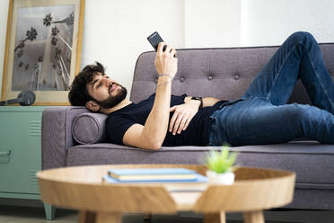 Lächelnder Mann, der sein Smartphone benutzt, während er sich auf dem Sofa im Wohnzimmer zu Hause entspannt - GIOF11976