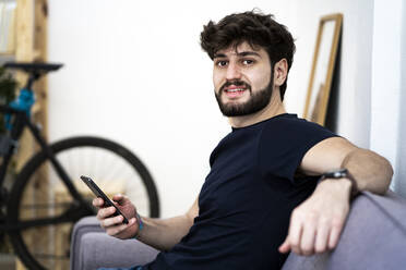 Junger Mann mit Smartphone sitzt zu Hause auf dem Sofa - GIOF11979