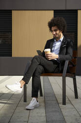 Afro man having coffee using smart phone while sitting on chair at street during sunny day - VEGF04088