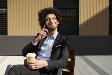 Lächelnder Afro-Mann, der mit seinem Smartphone telefoniert und eine Kaffeetasse hält, während er an einem sonnigen Tag auf einem Stuhl sitzt - VEGF04089