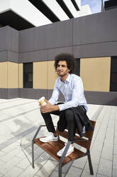 Man holding coffee cup while sitting on chair at street in city - VEGF04095