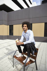 Mann hält Kaffeetasse, während er auf einem Stuhl auf der Straße in der Stadt sitzt - VEGF04095
