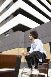 Afro man with coffee cup looking away while sitting on chair in city - VEGF04096