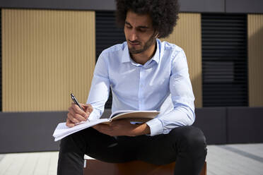 Afro man writing in book while sitting on chair at street - VEGF04098