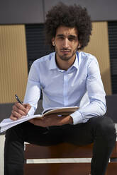 Mid adult man with pen and book sitting on chair at street - VEGF04099