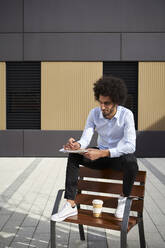 Mann mit Kaffeetasse, der auf einem Stuhl sitzend in ein Buch schreibt, während er an einem sonnigen Tag sitzt - VEGF04100