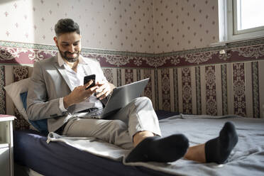 Smiling male professional using smart phone while sitting on bed at home - FBAF01653