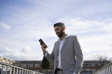 Smiling businessman using mobile phone in front of sky - FBAF01662