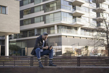 Smiling man using mobile phone while sitting on bench - FBAF01672