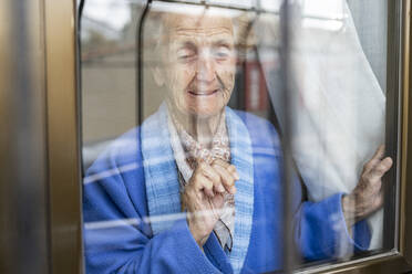 Senior woman looking through window of house - JCCMF01555