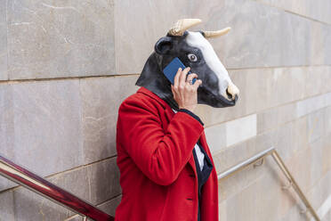 Businesswoman with bull mask talking on smart phone while standing near railing on wall - DLTSF01671
