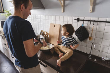 Father preparing food while looking at cute son sitting on kitchen counter at home - VYF00423