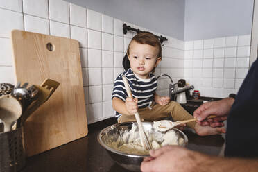 Verspieltes Kleinkind, das seinem Vater beim Mischen von Zutaten hilft, während es zu Hause auf dem Küchentisch sitzt - VYF00424