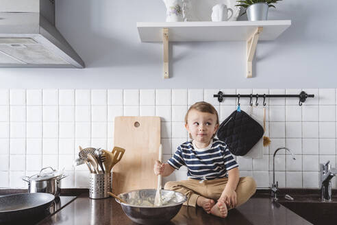 Verspieltes Kleinkind mit Schöpfkelle und Schüssel, das wegschaut, während es auf der Küchentheke zu Hause sitzt - VYF00427