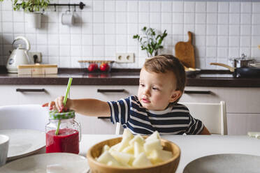 Niedlicher Junge hält Strohhalm in Einmachglas, während er am Esstisch in der Küche sitzt - VYF00437