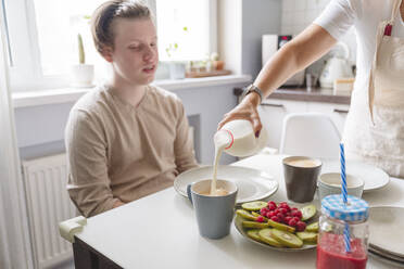 Frau gießt Milch in Tasse für Sohn im Teenageralter am Esstisch in der Küche - VYF00438
