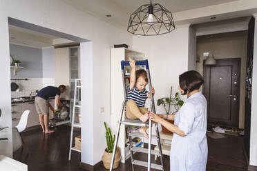 Mutter spielt mit ihrem Sohn auf der Leiter sitzend, während der Vater zu Hause in der Küche arbeitet - VYF00443