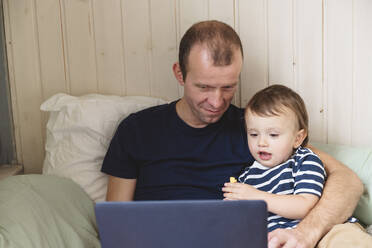 Mature man working on laptop while sitting with son on sofa at home office - VYF00449