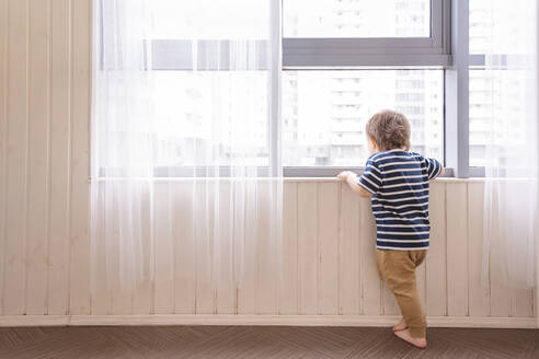 Junge schaut durch das Fenster, während er zu Hause steht - VYF00450