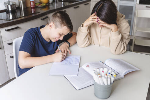 Frustrierte Mutter hilft ihrem Sohn bei den Hausaufgaben, während sie zu Hause am Esstisch sitzt - VYF00455