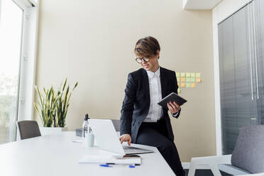 Geschäftsfrau, die einen Laptop benutzt und ein digitales Tablet am Schreibtisch im Büro hält - MEUF02195