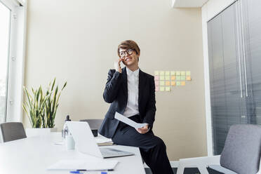 Smiling female professional looking away while talking on mobile phone at desk - MEUF02197