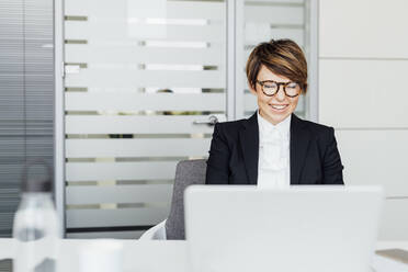 Glückliche Geschäftsfrau mit kurzen Haaren sitzt vor einem Laptop am Schreibtisch - MEUF02212