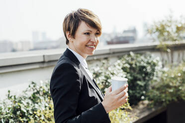 Smiling female entrepreneur holding disposable coffee cup while looking away - MEUF02215