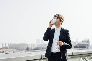 Weiblicher Geschäftsmann trinkt Kaffee, während er eine Brille an einer Stützmauer hält - MEUF02216