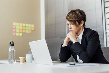 Nachdenkliche Unternehmerin mit Hand am Kinn, die auf ihren Laptop schaut, während sie im Büro sitzt - MEUF02218
