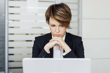 Nachdenkliche Unternehmerin mit braunem Haar, die im Büro vor einem Laptop sitzt - MEUF02220