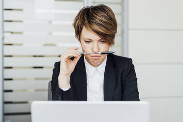 Businesswoman holding pen on lips while looking at laptop in office - MEUF02221