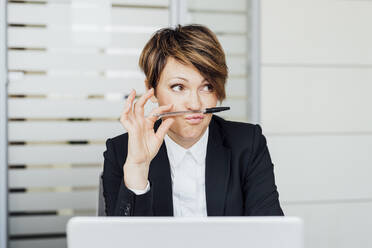 Geschäftsfrau schaut weg, während sie vor einem Laptop im Büro einen Stift auf den Lippen hält - MEUF02223