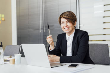 Smiling female entrepreneur holding pen in front of laptop at desk in office - MEUF02224