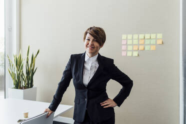 Smiling female entrepreneur with hand on hip standing at desk in office - MEUF02236