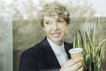 Lächelnde Geschäftsfrau, die wegschaut, während sie eine Einweg-Kaffeetasse hält, gesehen durch ein Glasfenster - MEUF02244
