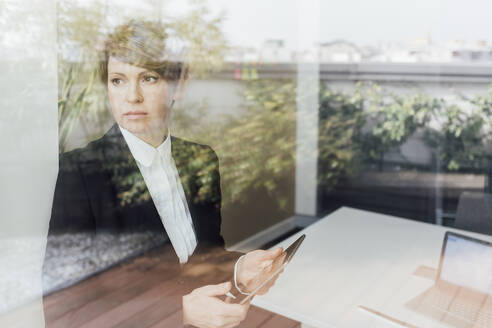 Serious businesswoman holding mobile phone while looking through window in office - MEUF02245