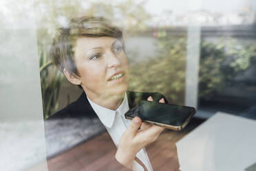 Zufriedene Geschäftsfrau, die ihr Handy in der Hand hält und durch ein Fenster im Büro schaut - MEUF02249