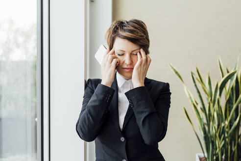 Müde Geschäftsfrau mit geschlossenen Augen hält Handy im Büro - MEUF02255