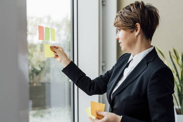 Female professional sticking adhesive notes on glass wall in office - MEUF02262