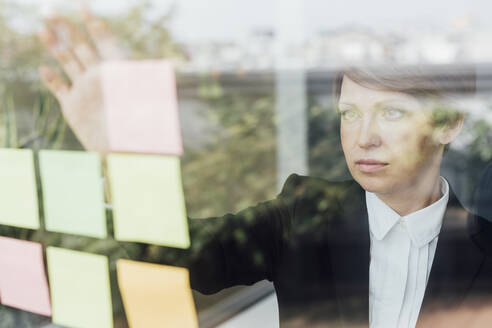 Serious businesswoman sticking adhesive notes seen through glass wall in office - MEUF02263