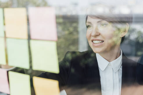 Smiling female entrepreneur looking at adhesive notes on glass wall - MEUF02264