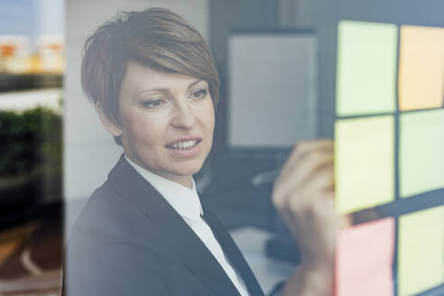 Female business professional writing on adhesive notes in office seen through glass wall - MEUF02265