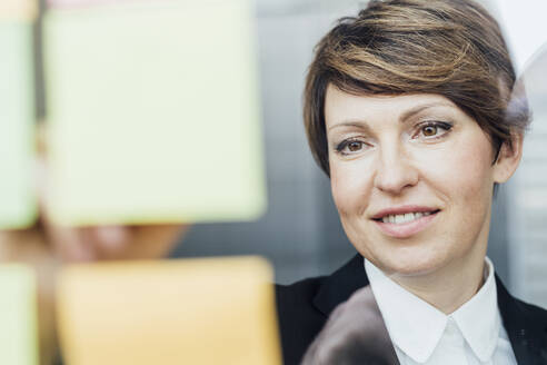 Contented businesswoman looking at adhesive notes sticked on glass wall in office - MEUF02266