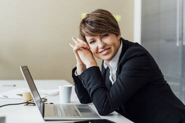 Happy businesswoman with hands clasped sitting in front of laptop at desk in office - MEUF02272